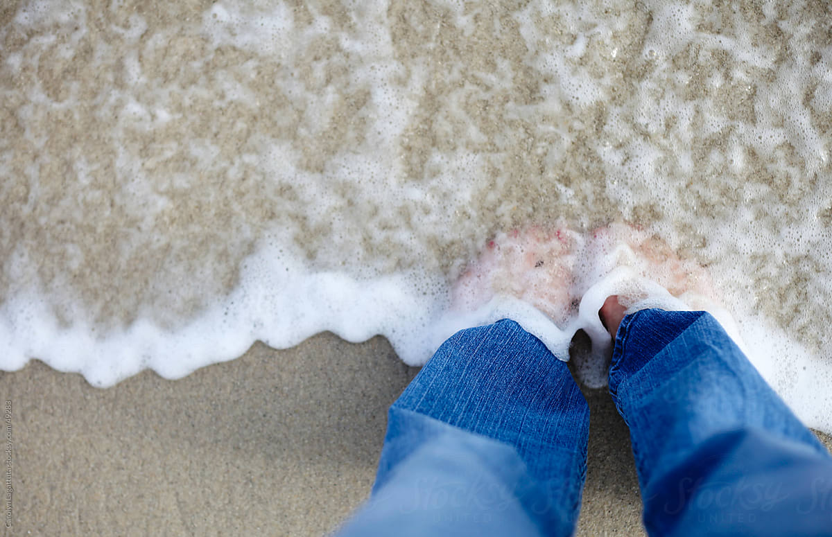 Girl standing in the surf with jeans on and the tide is washing over her feet.