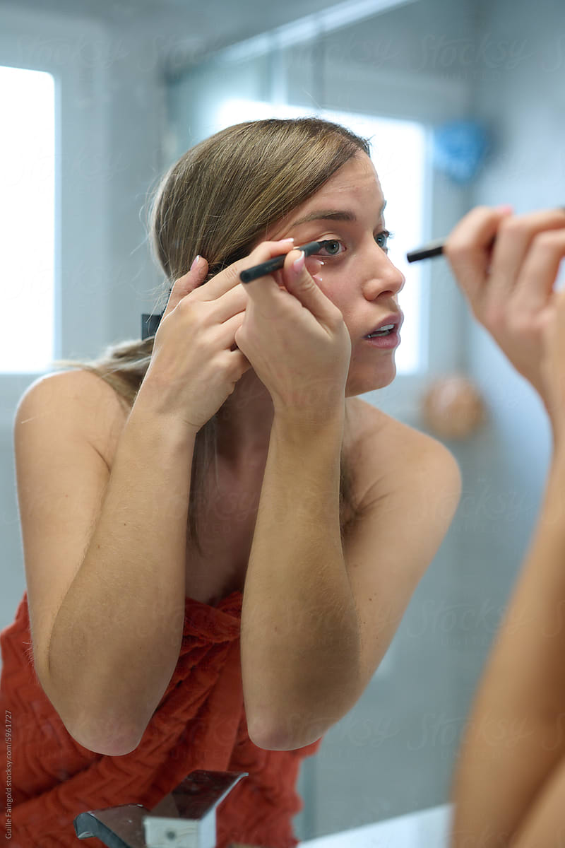 Woman applying eyeliner in front of bathroom mirror.