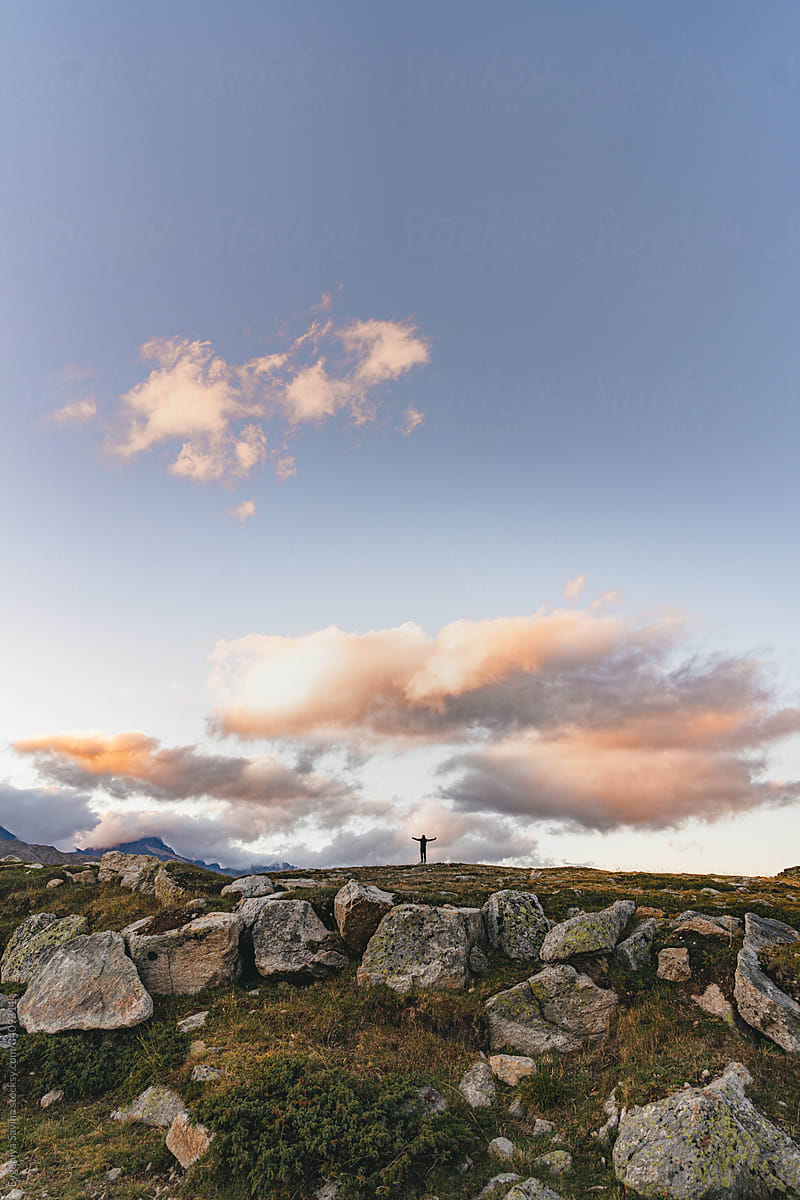 A figure of a man standing on a big rock in the mountains with sunset clouds on the background.