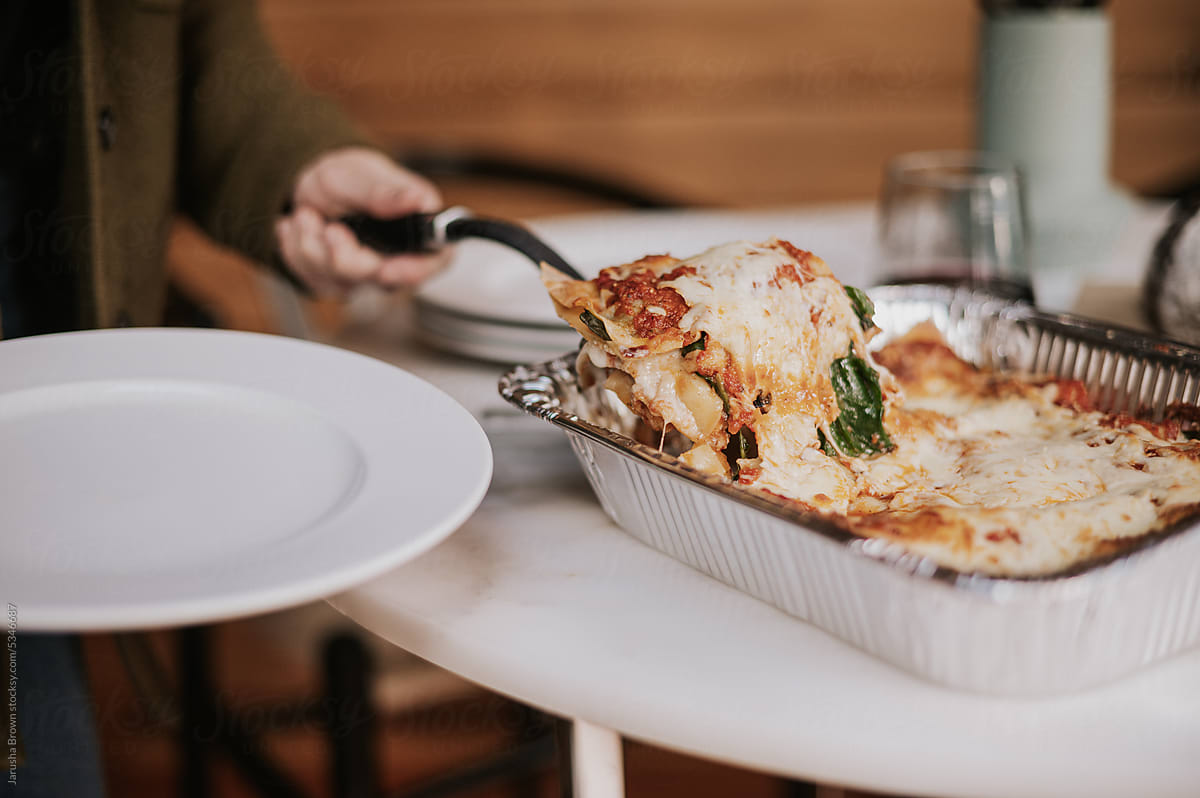 Woman serves a portion cooked lasagna from a tray.