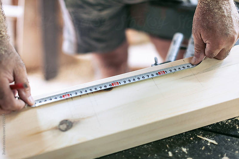 A close up of a carpenters hands measuring a piece of wood using a tapeline and a pencil