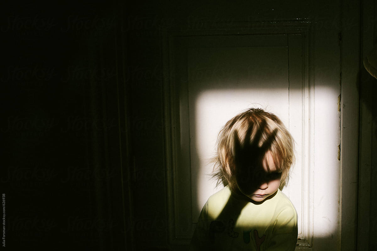 Small child in patch of light with shadow of adult hand over face.