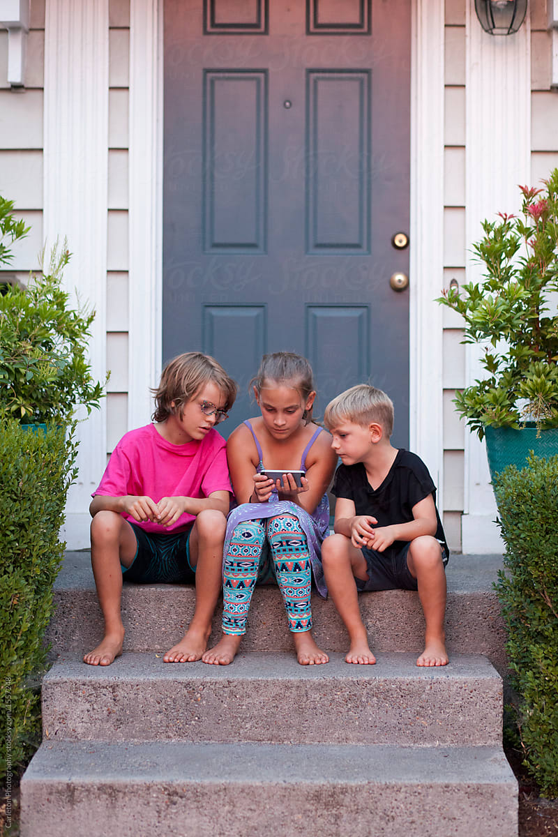 Three kids sitting on a front doorstep looking at a smartphone