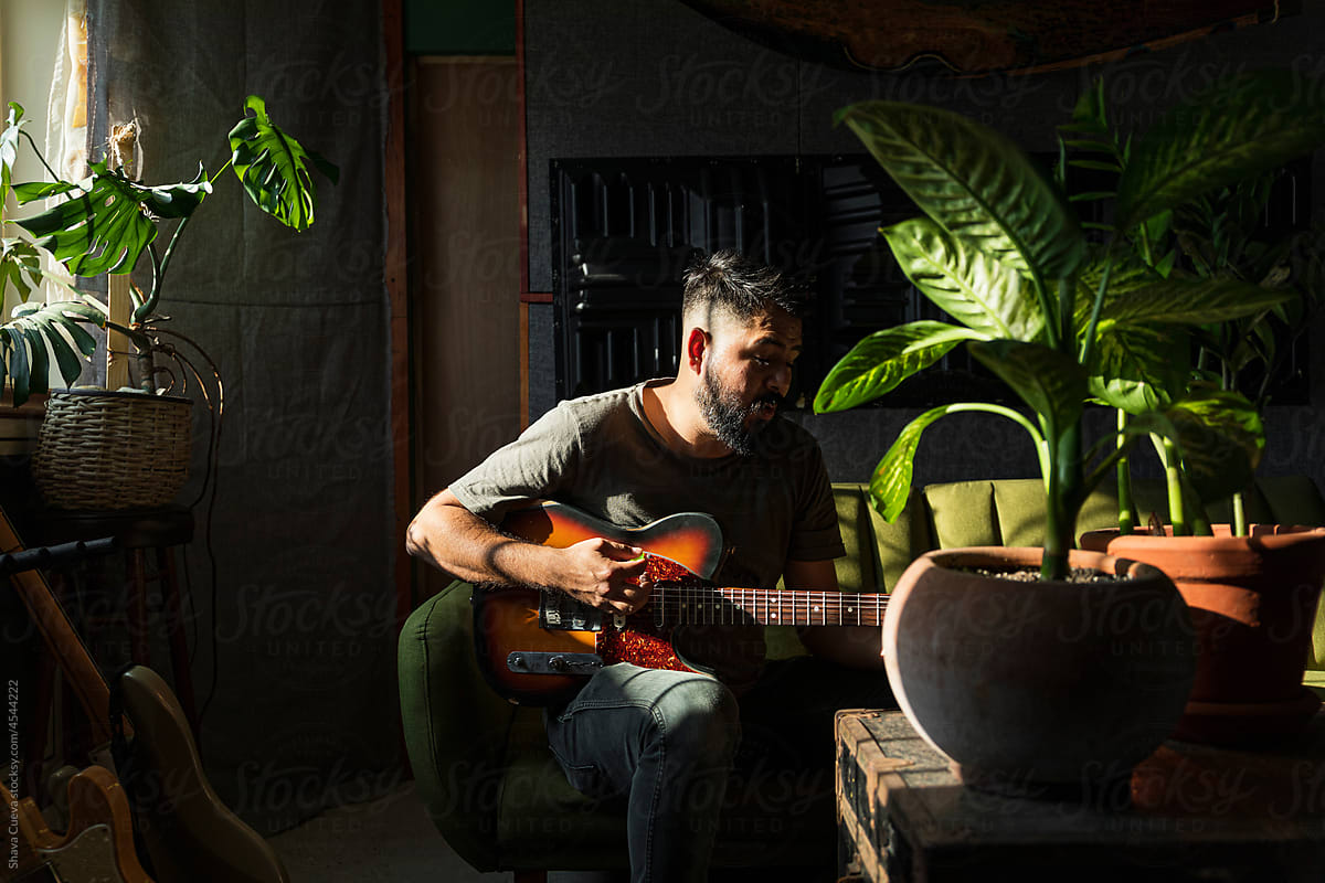 Man with short hair playing an electric guitar in a room with plants