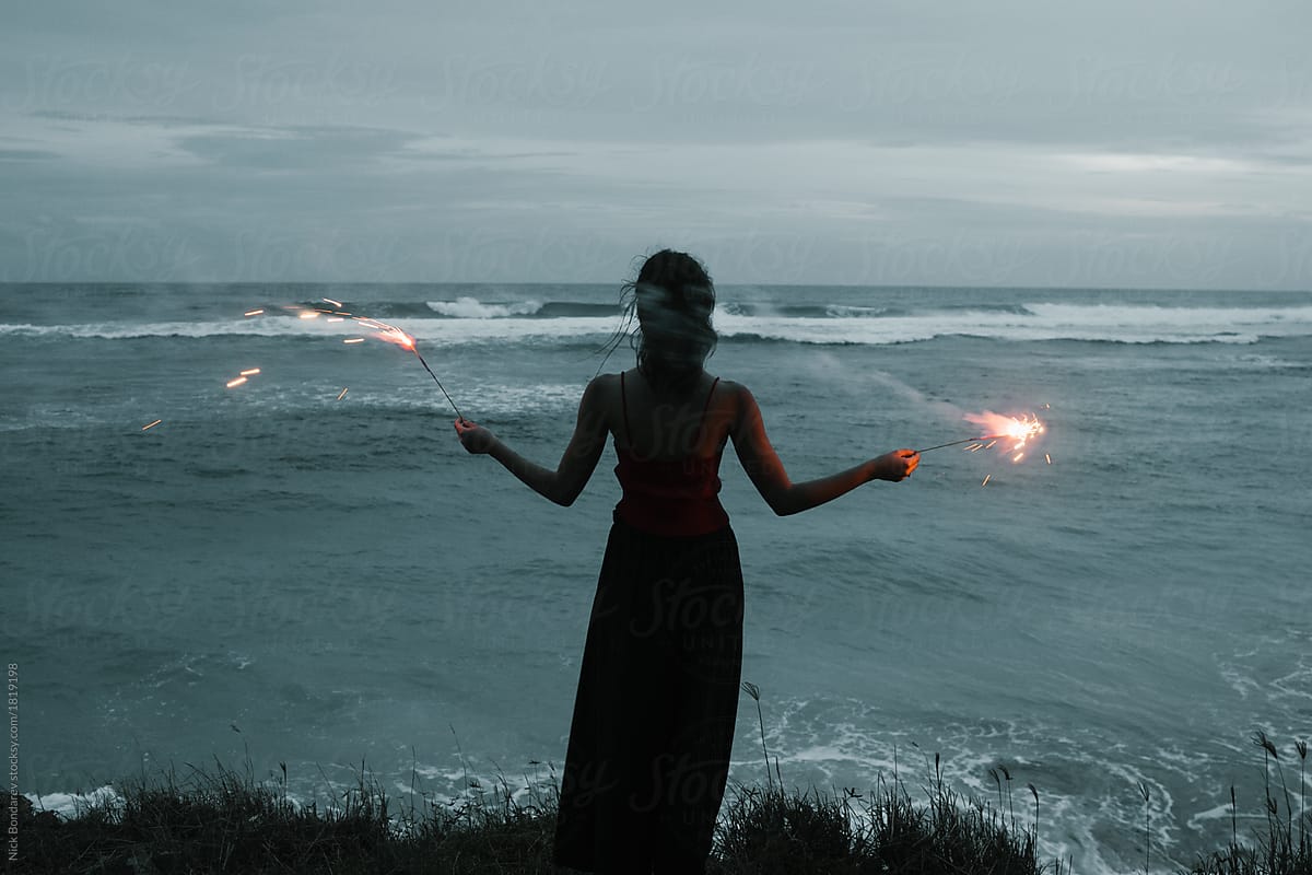 young fashion woman stand on the ocean coast with fireworks sparkles in hands with sea view