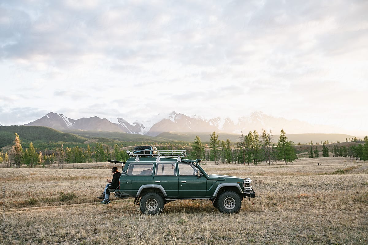 Two anonymous men sitting on the green jeep parked in the wild area surrounded by mountains