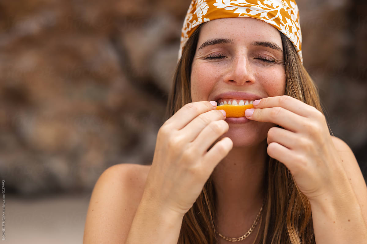 Content woman in bandana eating orange on coast