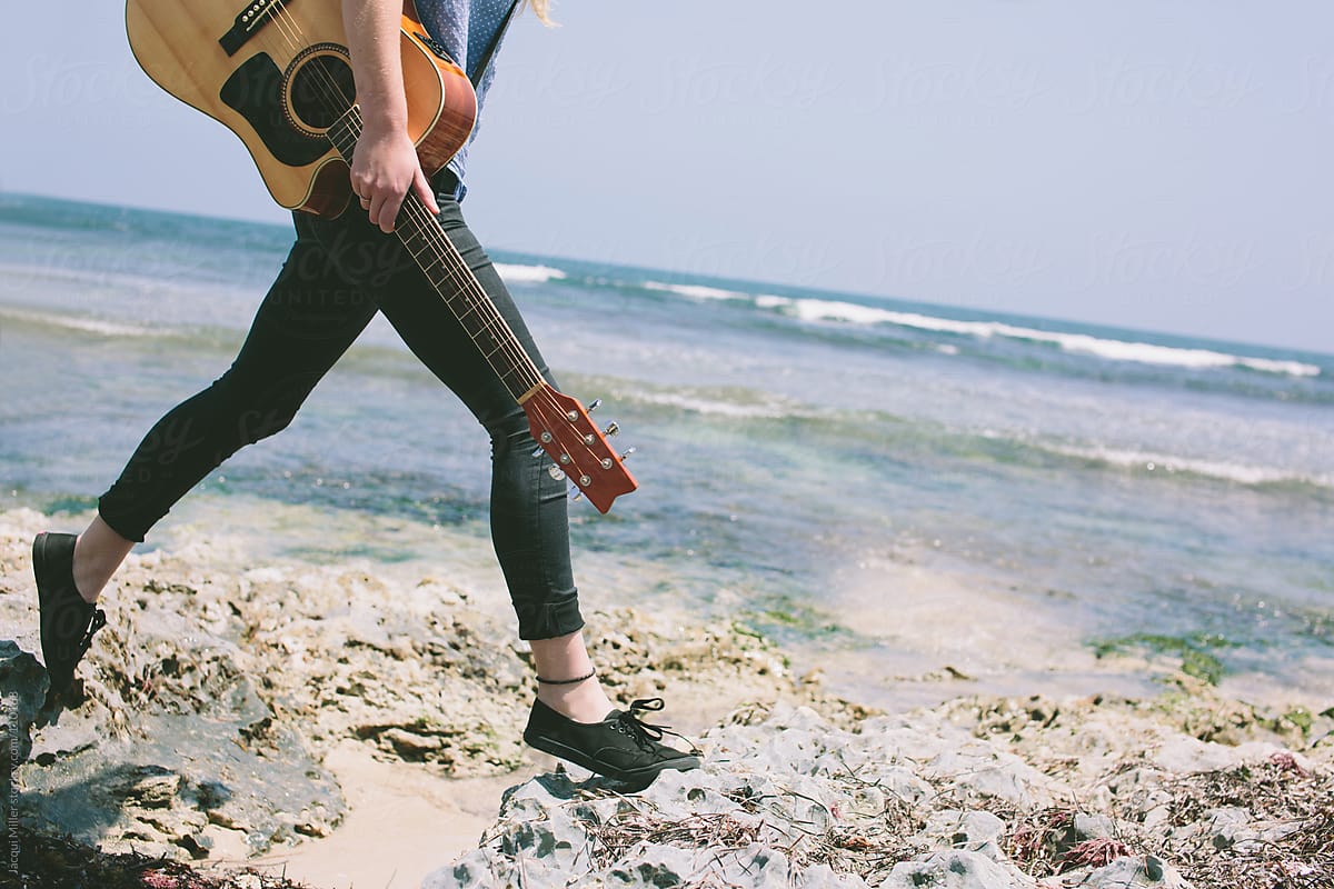 Girl walking along rocks at the beach with her guitar