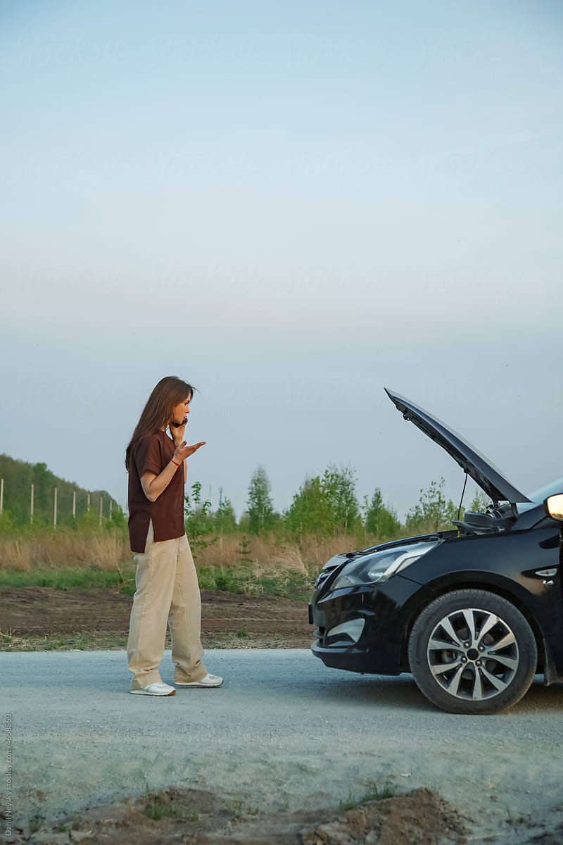Puzzled woman talking on smartphone near broken car