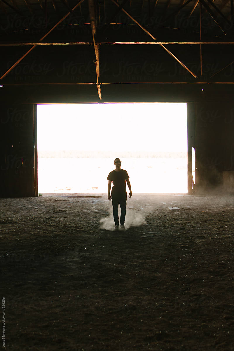 Young man walking through empty barn kicking up dirt floor with bright white behind silhouette