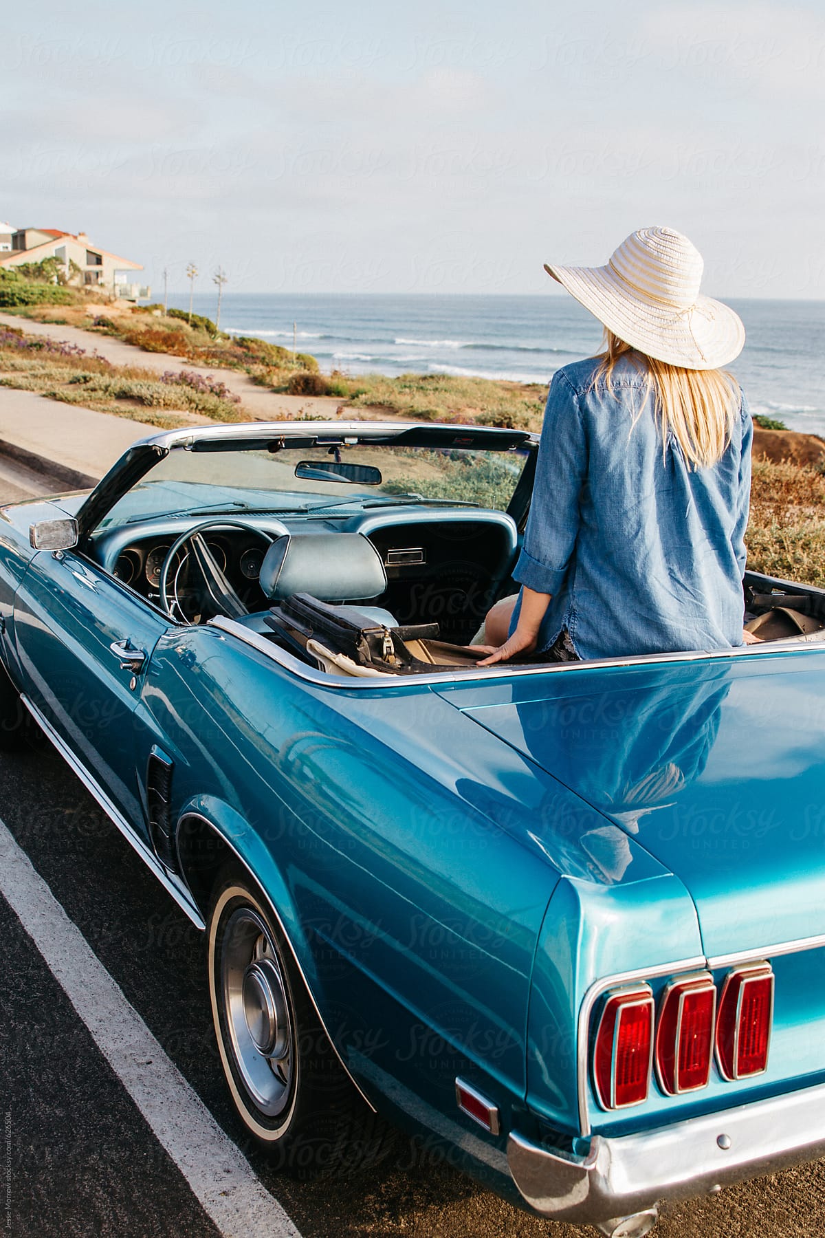 young woman with sun hat sitting on back of blue convertible car by ocean