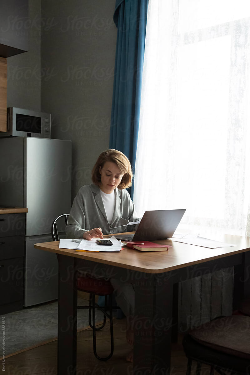 Focused woman working on laptop at home while using smartphone