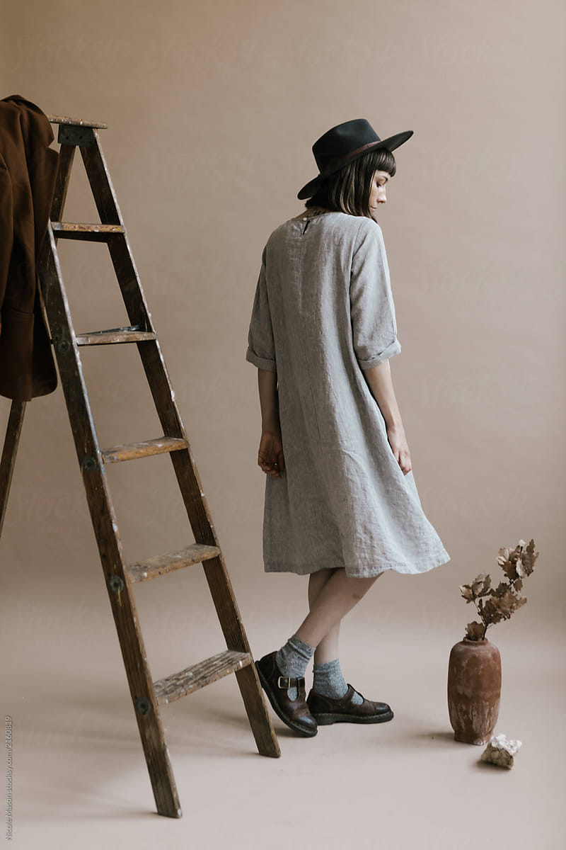 woman in grey dress and hat posing next to wooden ladder in photo studio