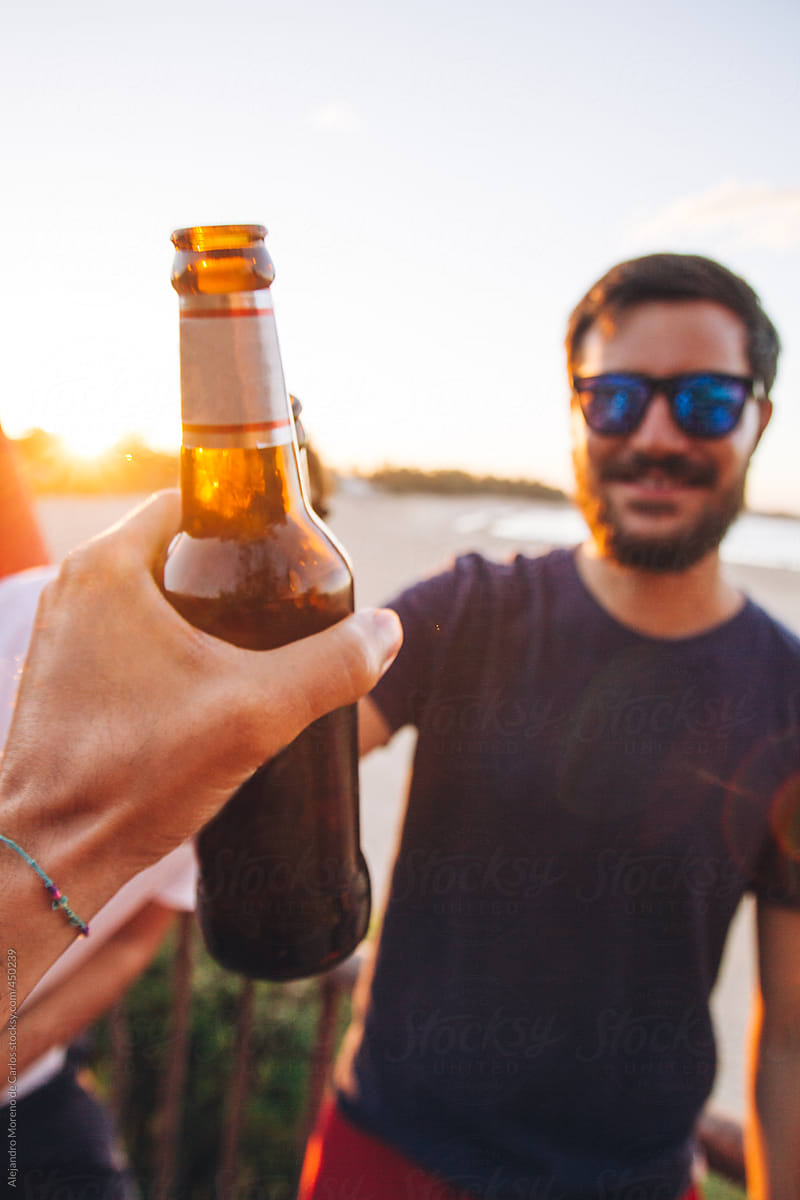 POV shot of a man toasting a bottle of beer with a friend on the beach at sunset