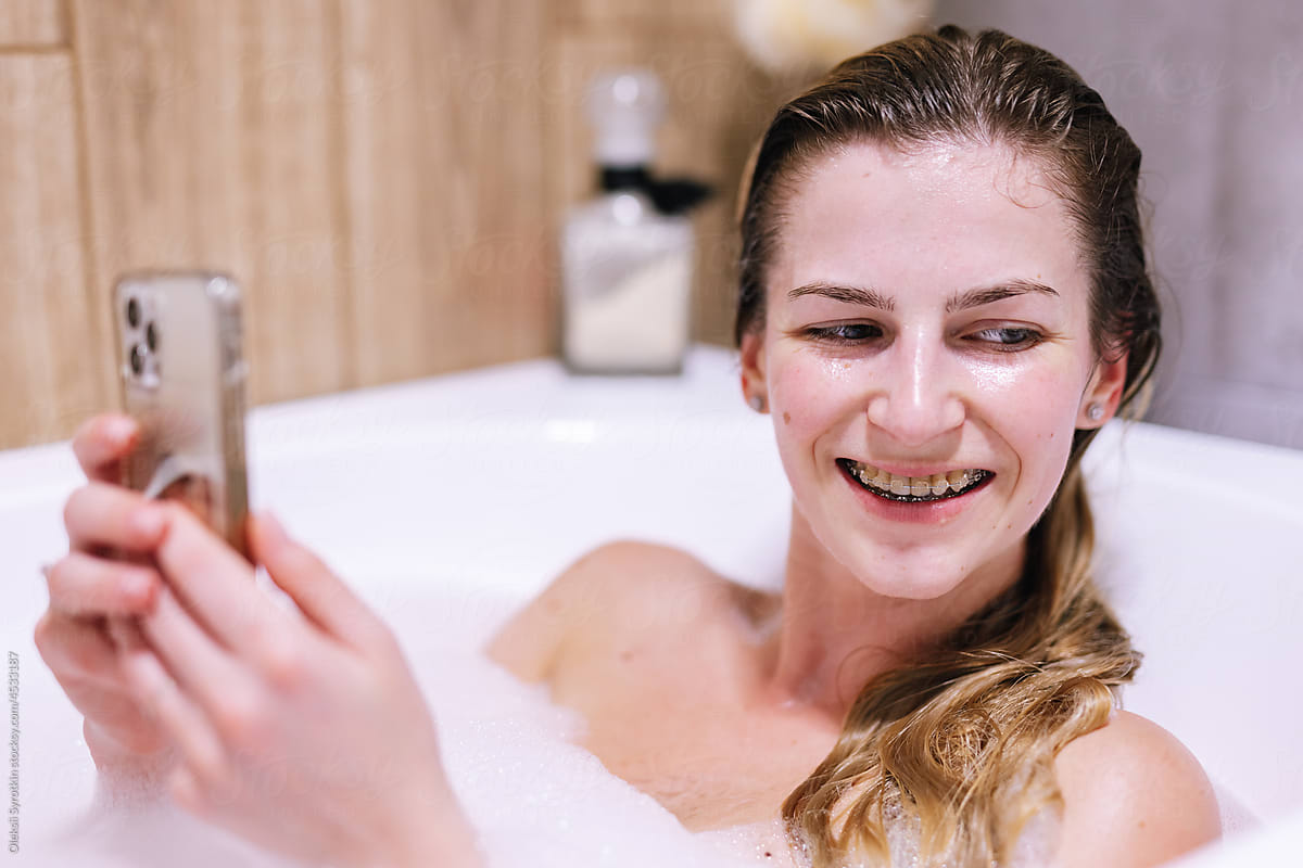 Blonde girl holding smartphone in bath with foam and smiling