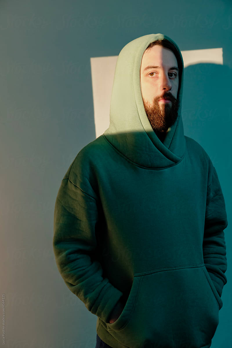 A man in a hood and a green sweatshirt stands against a lighted wall