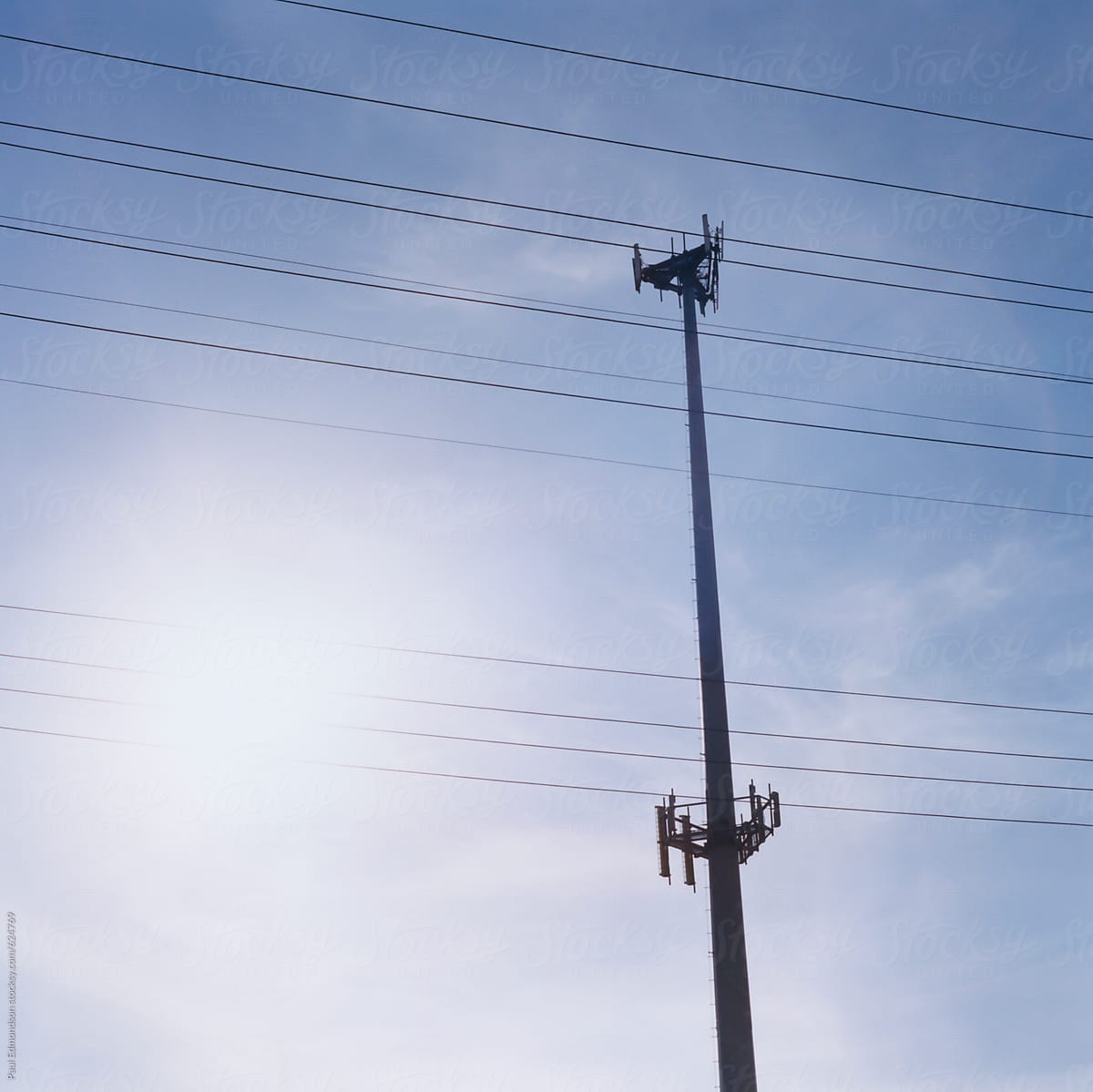 Cellular phone tower and utility wires across sky, bright sun in distance