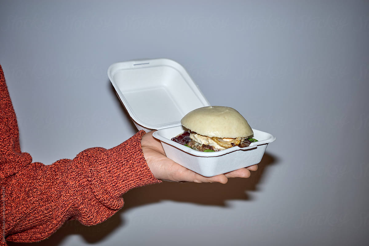 Hand holding burger in styrofoam container against grey background