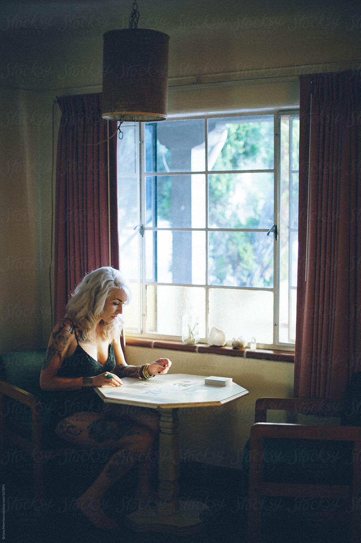 Blond Woman with Tattoos Reading Tarot next to A Window