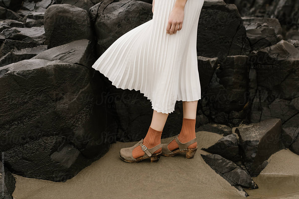 young person wearing flowing white skirt and orange socks standing in front of dark rocks