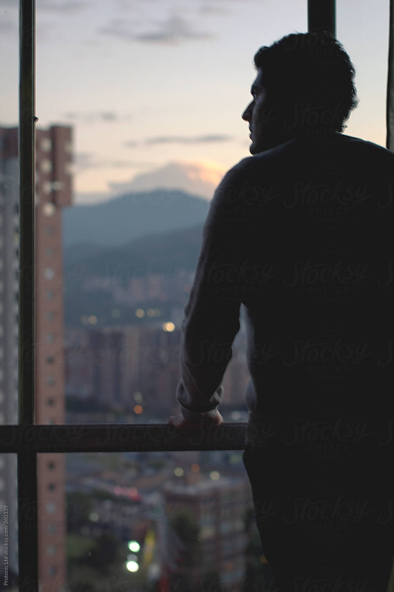 Silhouette of young man looking out from window in highrise with a view of a big city