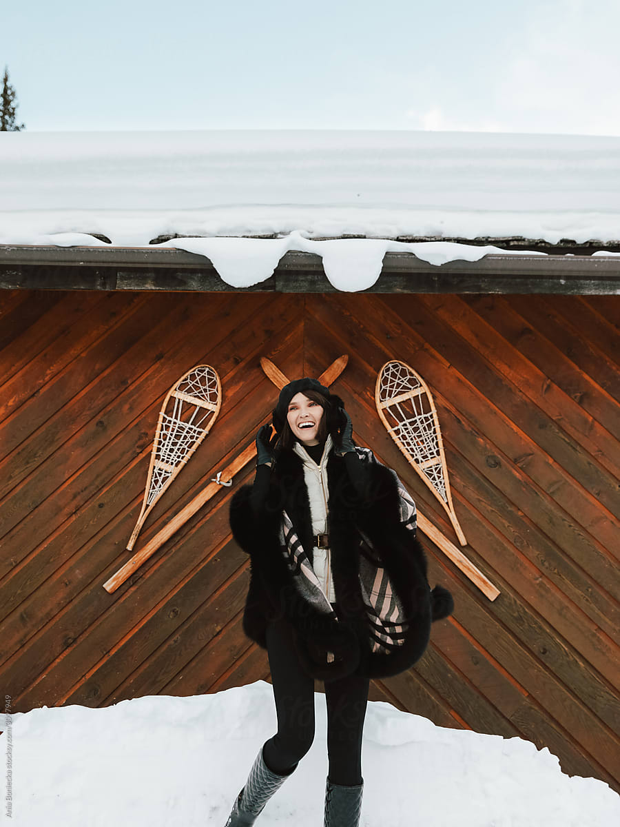 Stylish woman stands in front of decorative wood wall with snowshoes and wooden skis.