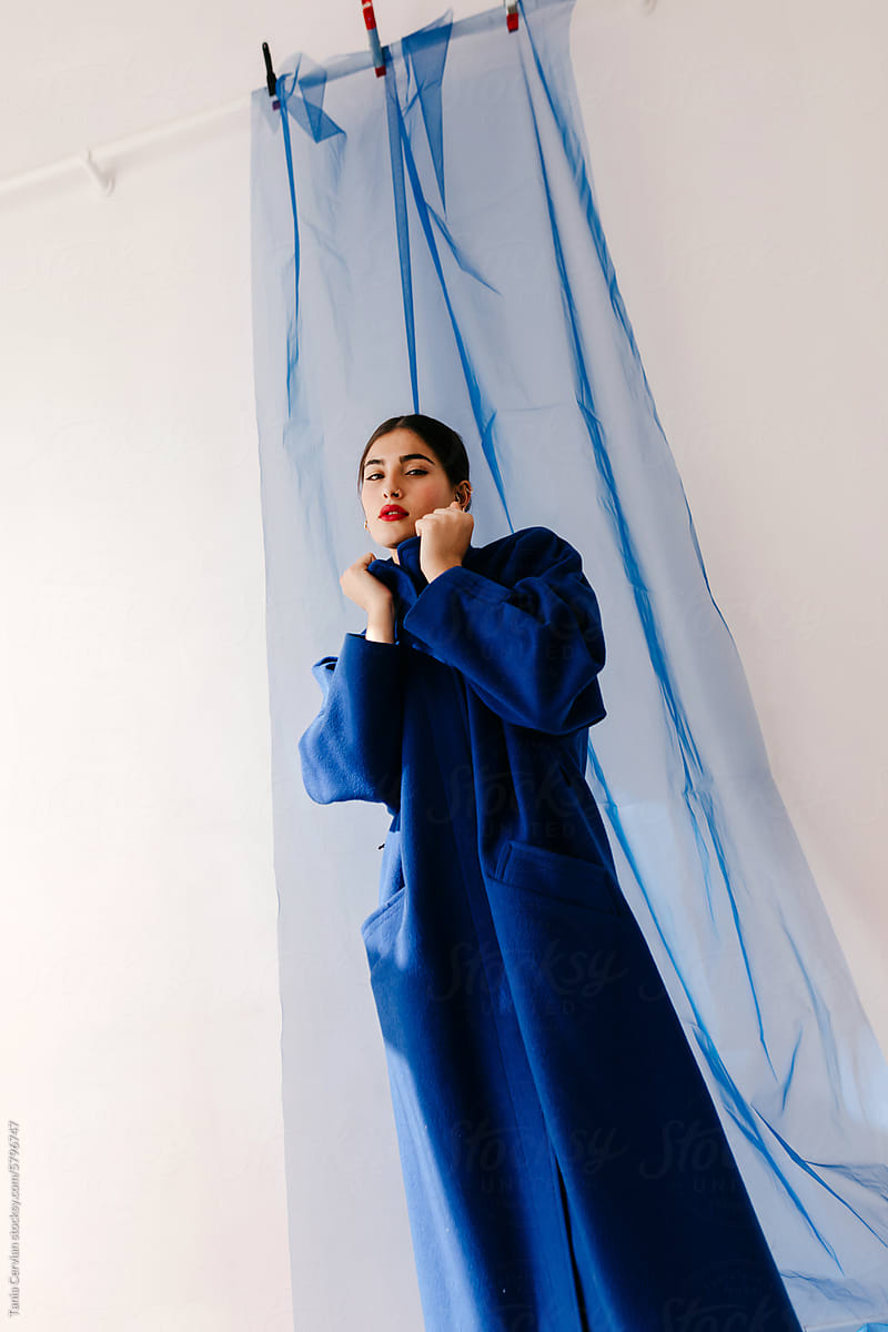 Elegant young woman in blue coat standing opposite blue fabric