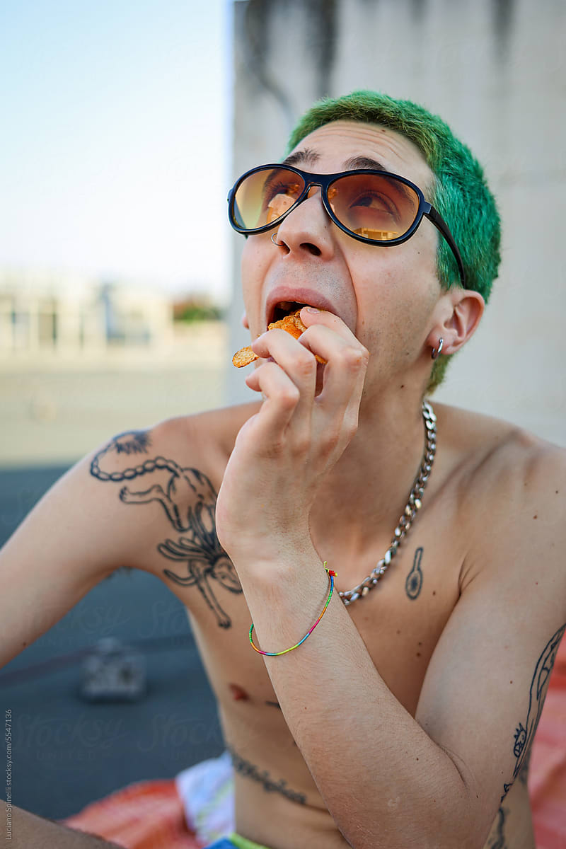 Tattooed shirtless guy with green hair and sunglasses eating chips