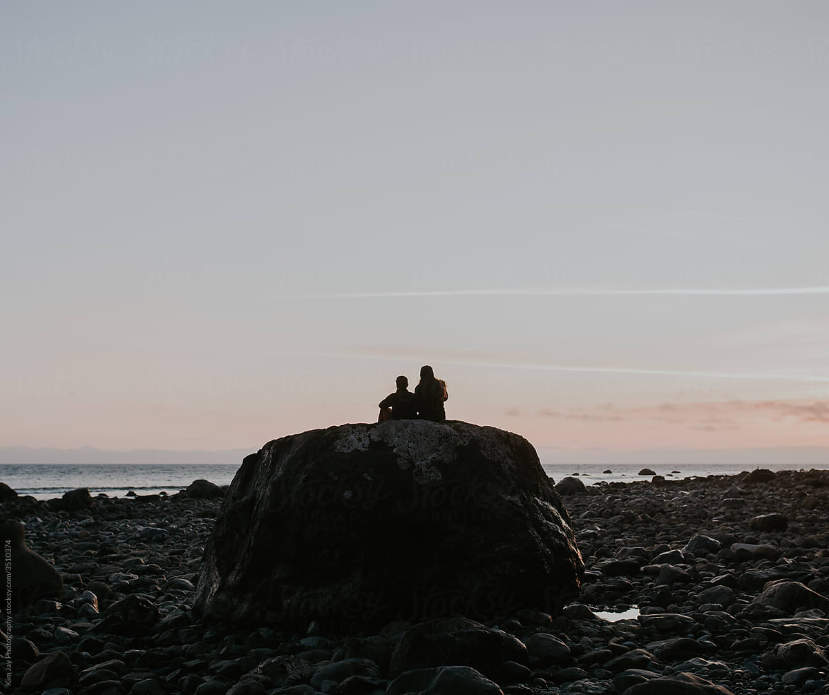 Two people sit on a large rock on the shore