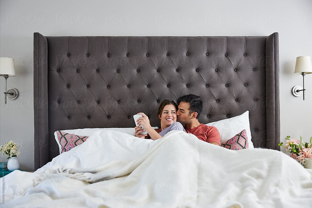 Young Millennial Couple In Bed Cuddling And Texting On A Cell Phone By Stocksy Contributor