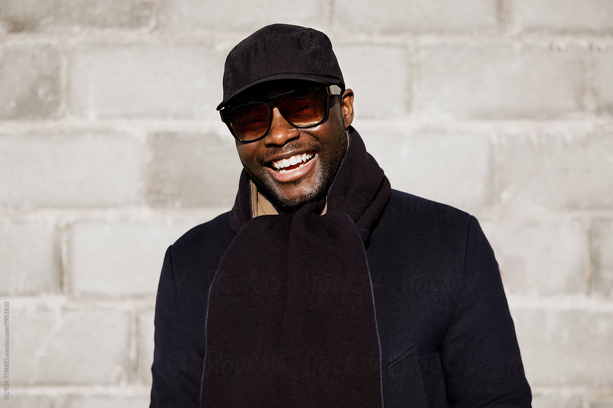 Smiling Stylish Man in Black Coat and Cap Against a Wall