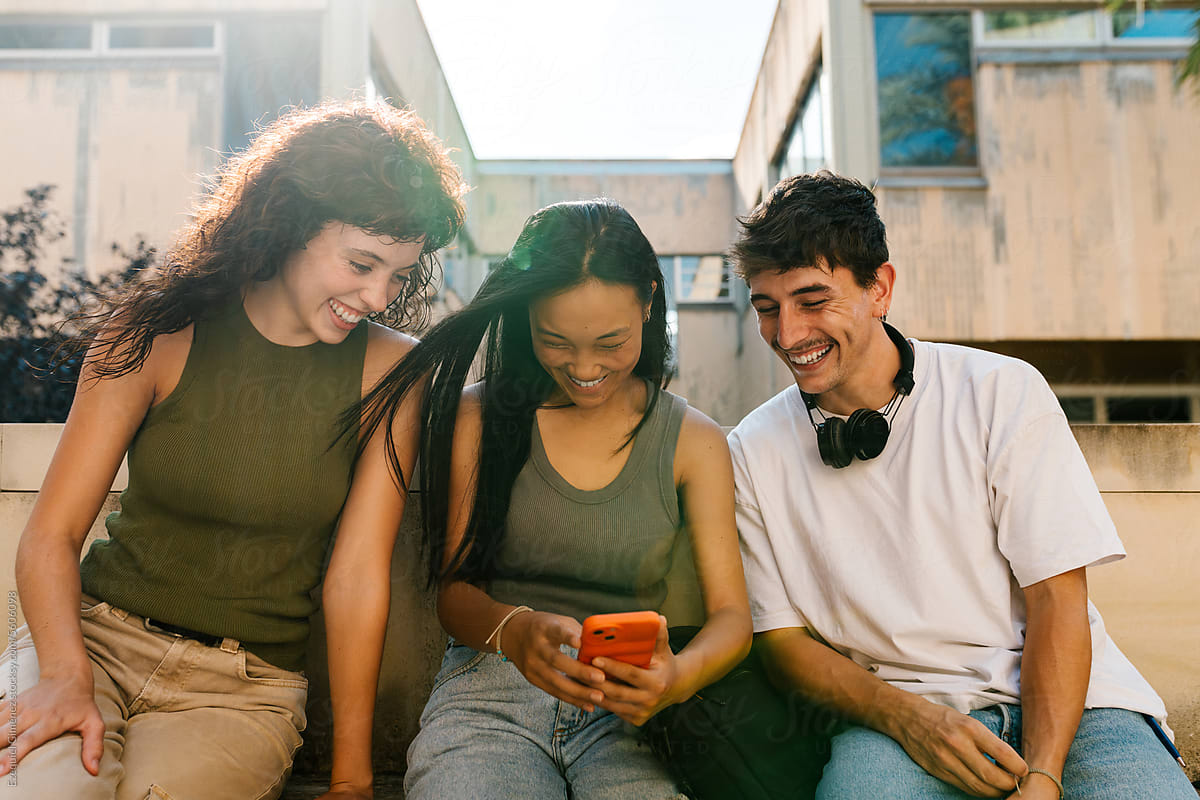 Smiling multi-ethnic students glance at their smartphone by day