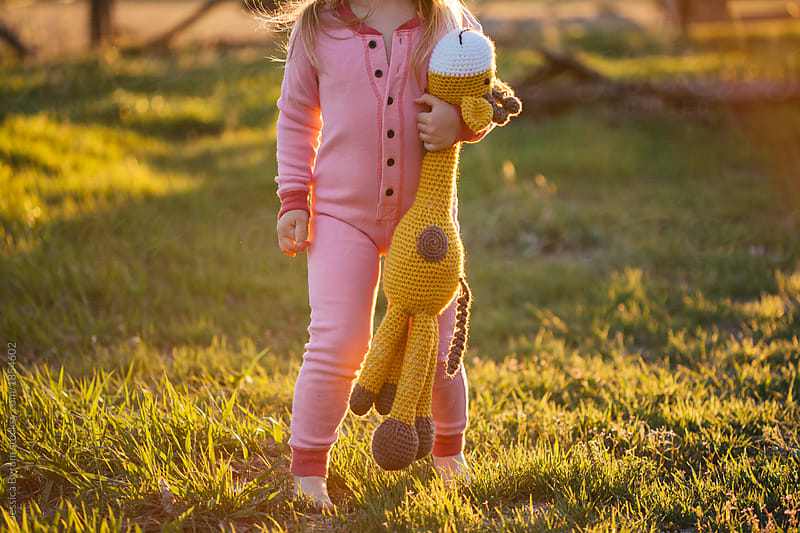 Toddler girl in pink pajamas walking with stuffed giraffe at sunset.