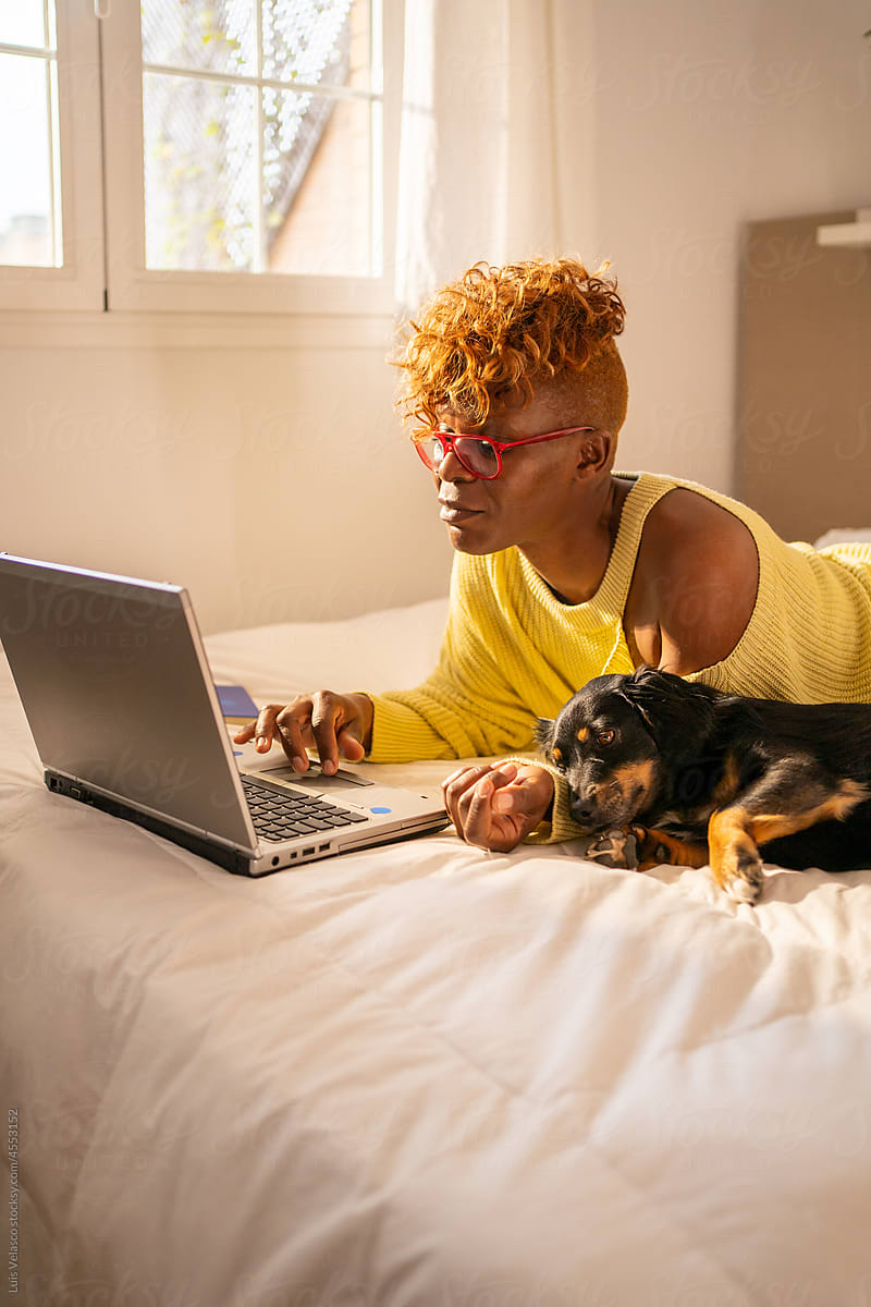 Cool Woman With Laptop Working From Home Next To Her Dog On The Bed.