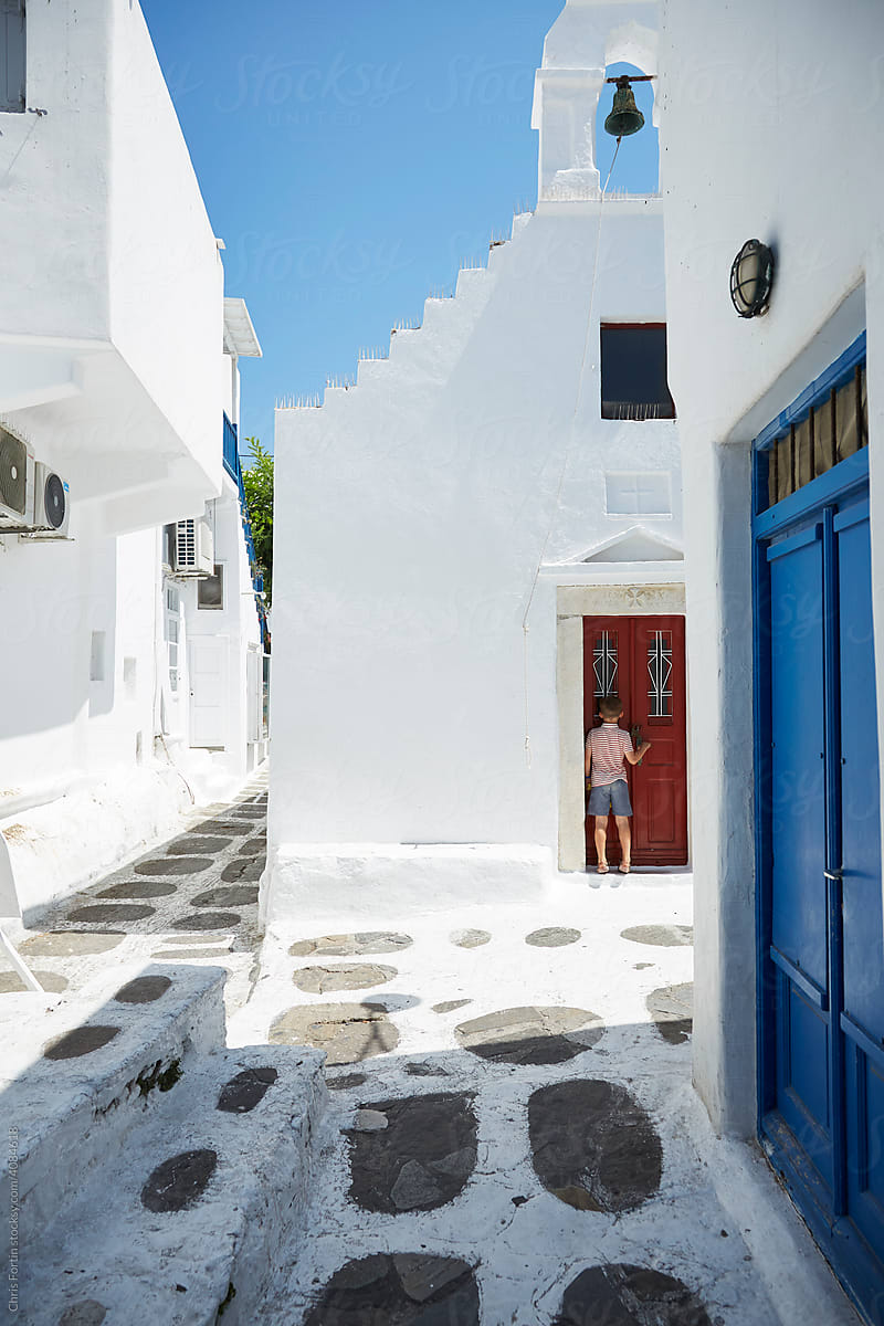 A quiet alleyway in Mykonos Greece, a young boy in striped shirt peers into the window of a small closed church with a red door