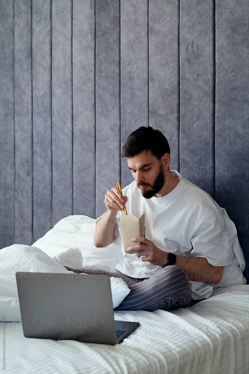 Calm man eating noodles and using laptop on comfy bed