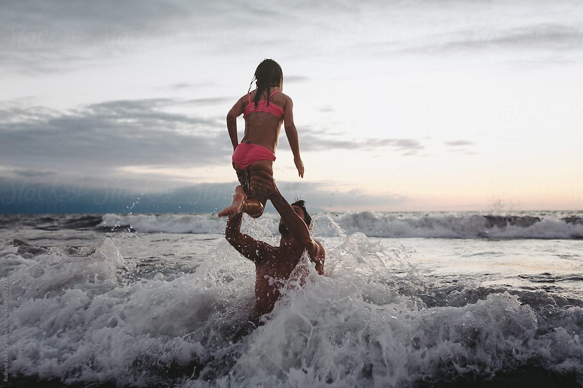 Young father lifting up little girl in ocean wave at sunset