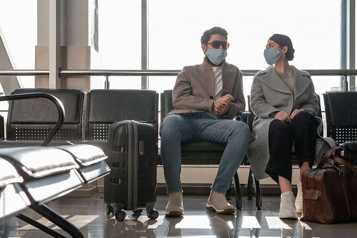 Couple in masks talking against window in airport waiting room