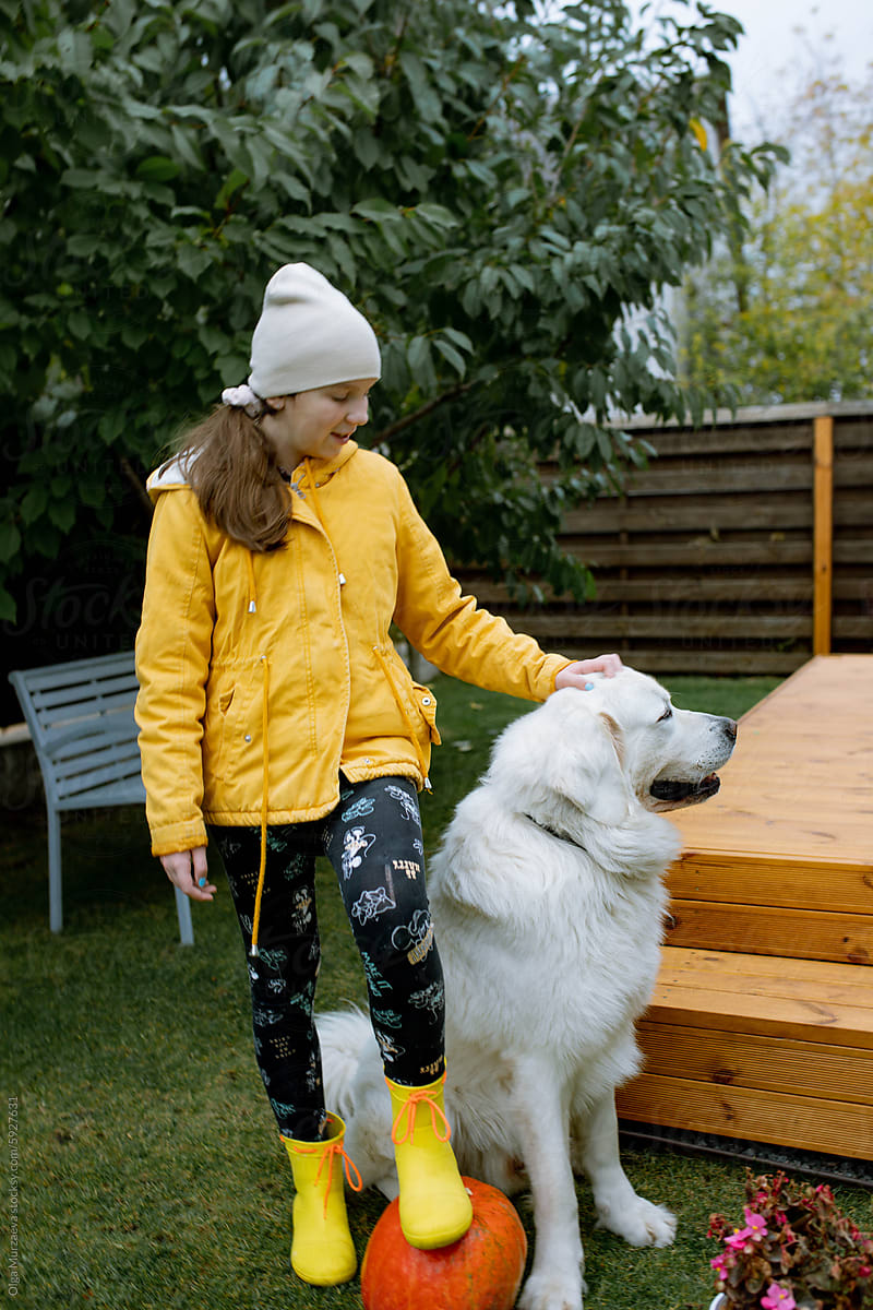 A girl in an orange jacket sits on a wooden terrace with a dog