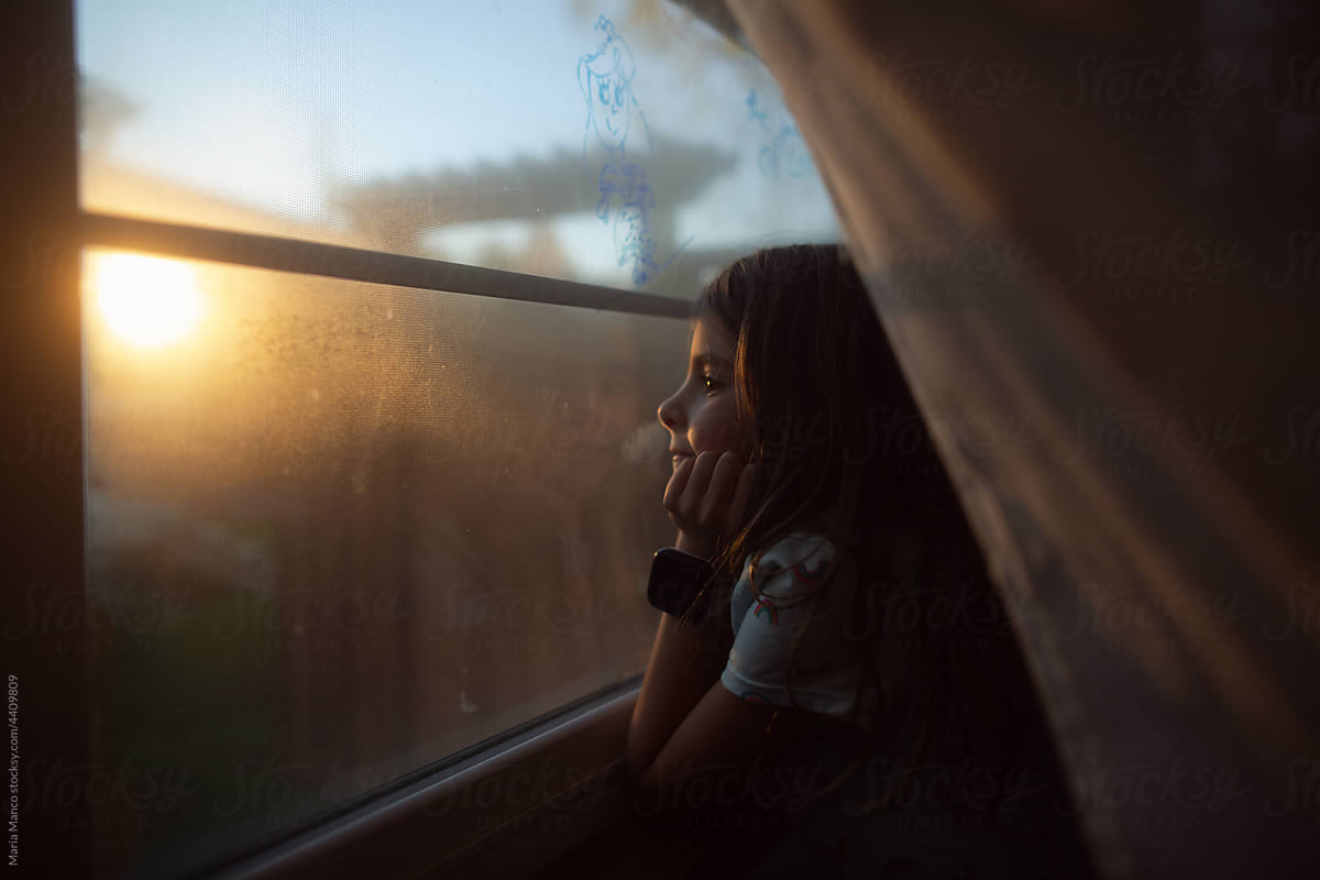 Young girls looks out window with sun setting outside