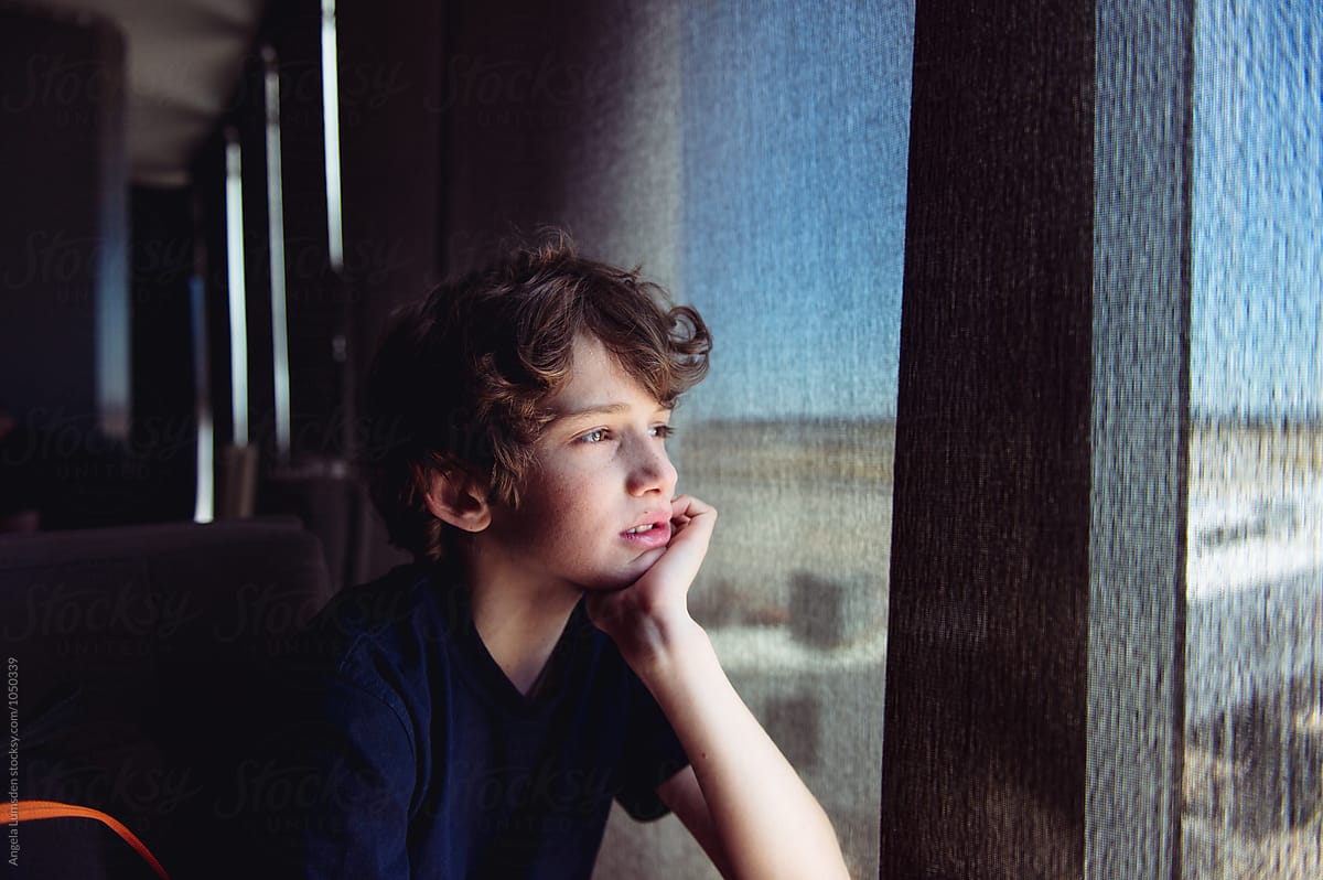 Teenage boy looks through the blinds of a large shaded window at an airport