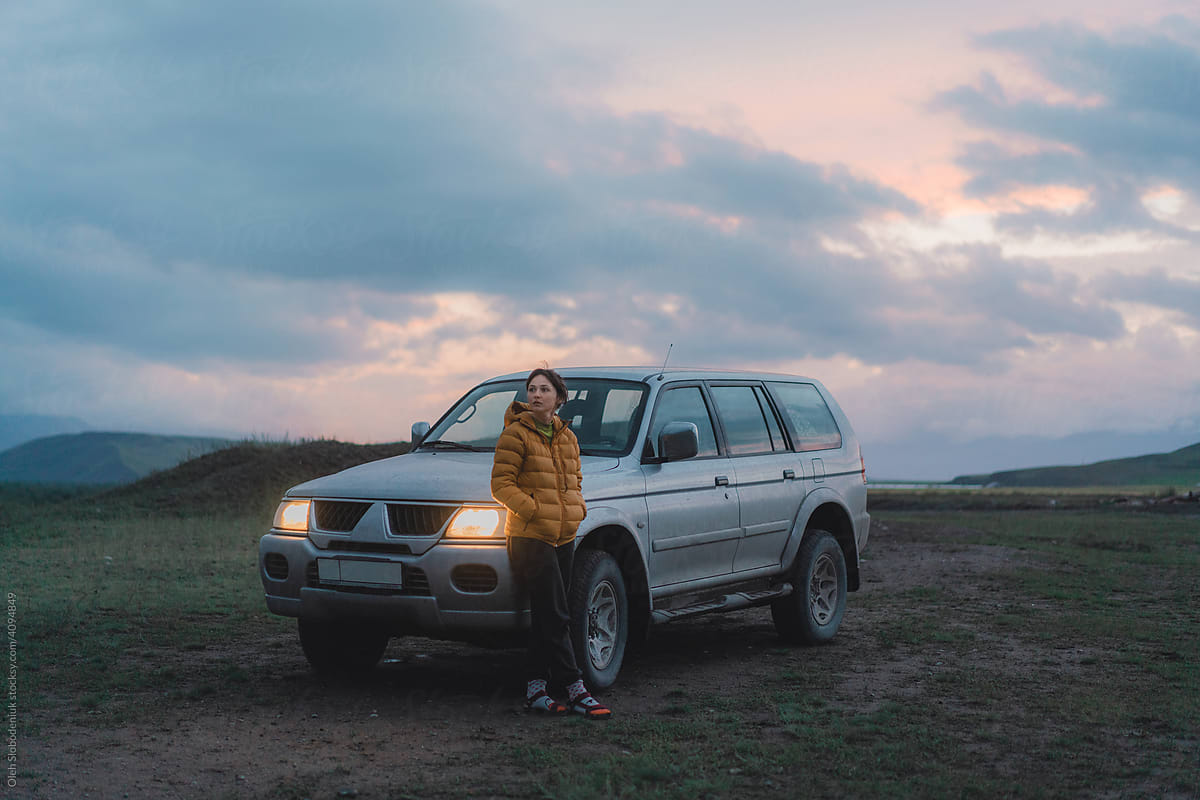 Woman standing near SUV car on dirt road at sunset