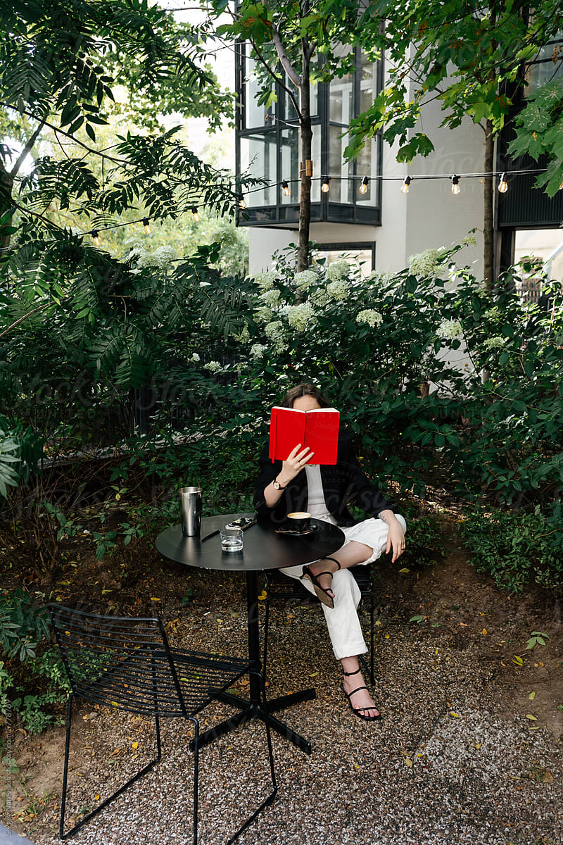 Woman covering face with book sitting at outdoor cafe table