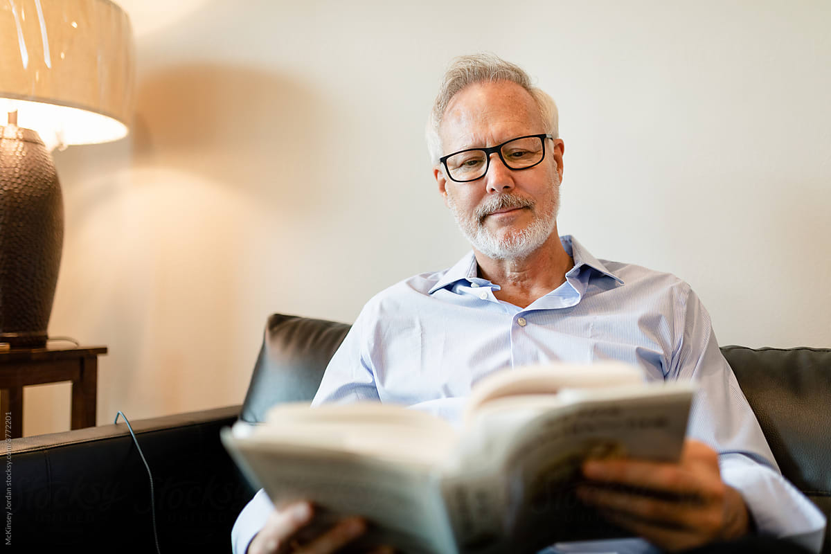 Older Man Wearing Glasses Sits on His Couch and Reads a Book