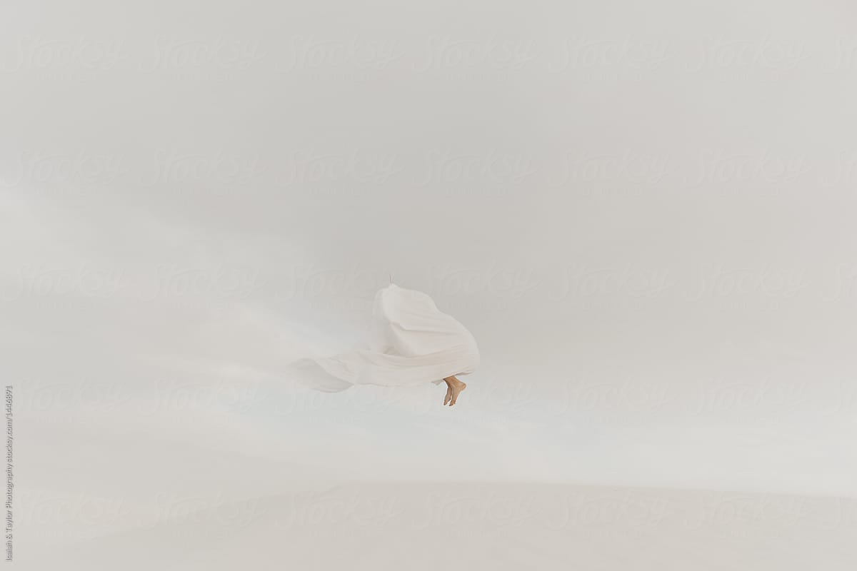 A camouflaged person jumping high in the desert while hiding under a ghost white sheet