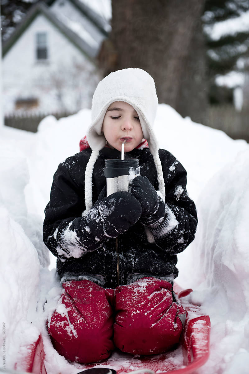 Boy sits in snowbank in full winter gear, drinking hot chocolate
