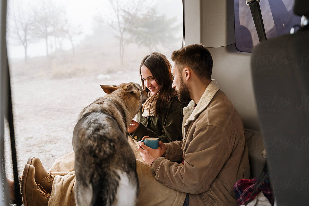 Young couple in a camper van in nature during winter having warm coffe with dog