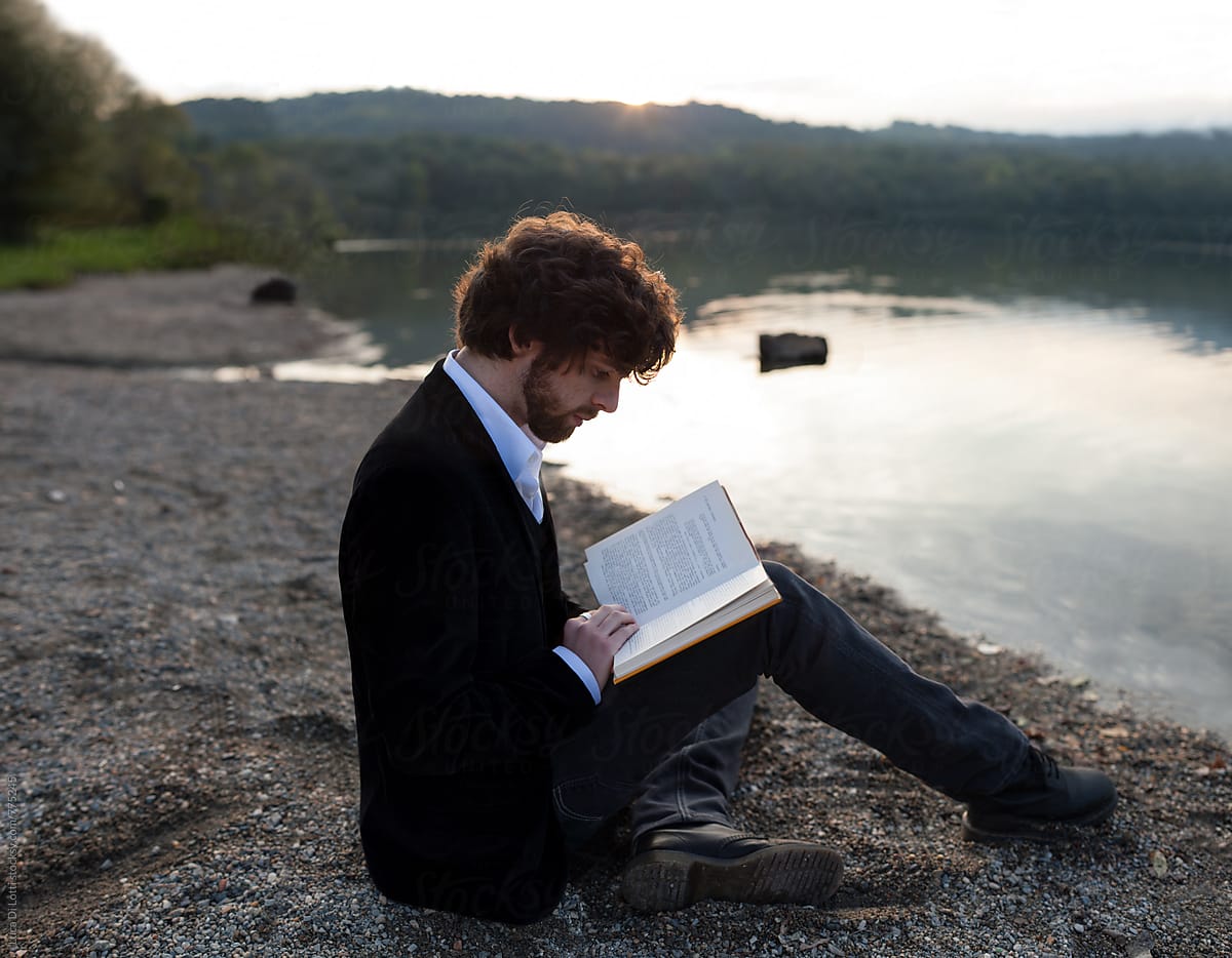 Young handsome bearded man reading a book sitting by a lake during sunset.