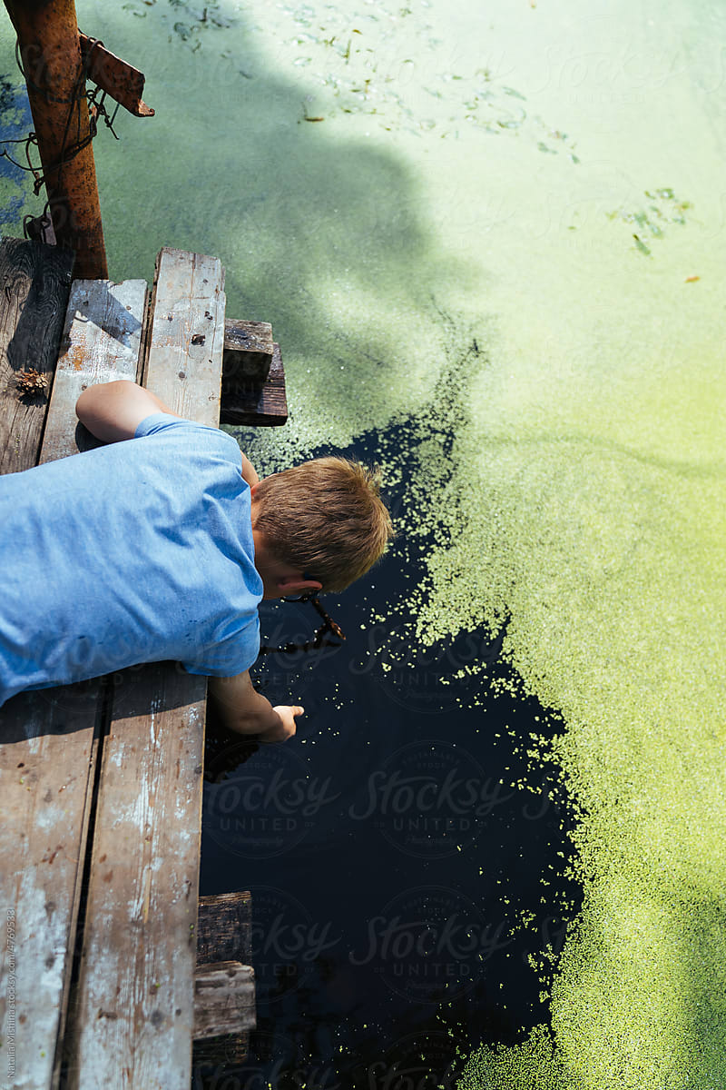 A boy on a wooden platform playing with water, duckweed on the pond.