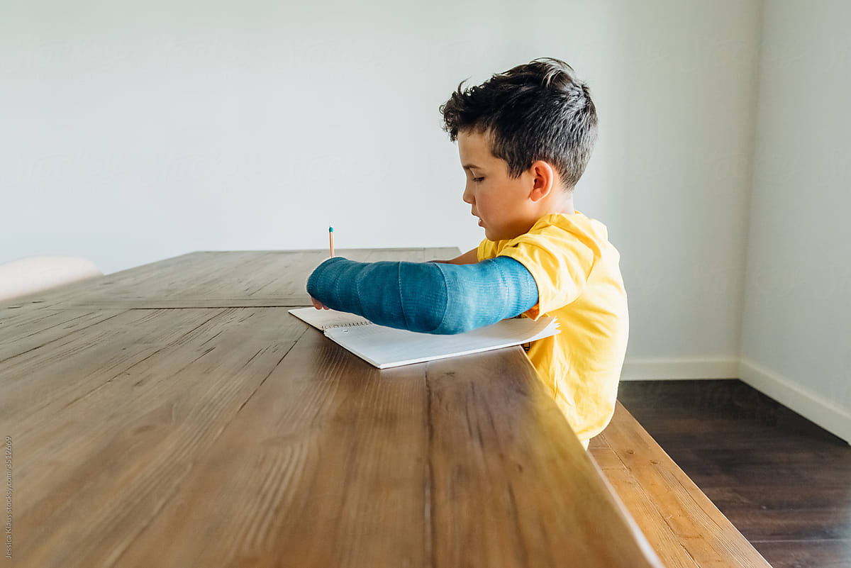 Boy with a blue cast doing schoolwork at the dining room table.